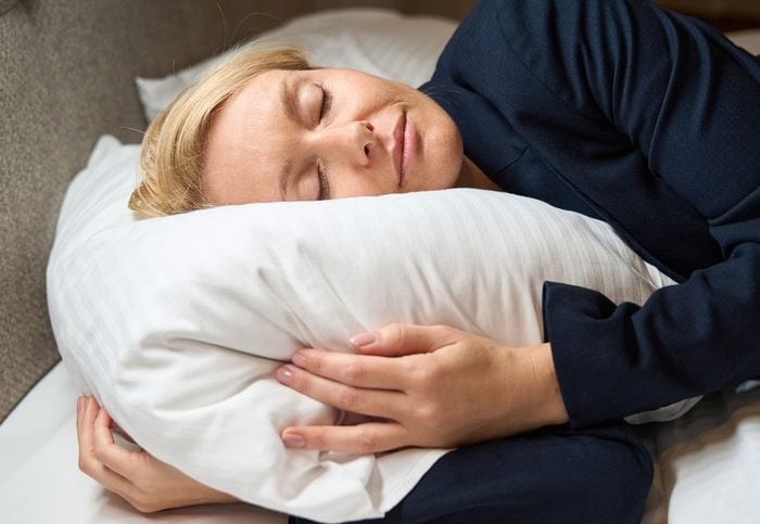 Žena spí na bielom vankúši v pohodlnom oblečení, ruky jemne objímajú vankúš, pokojný výraz tváre.