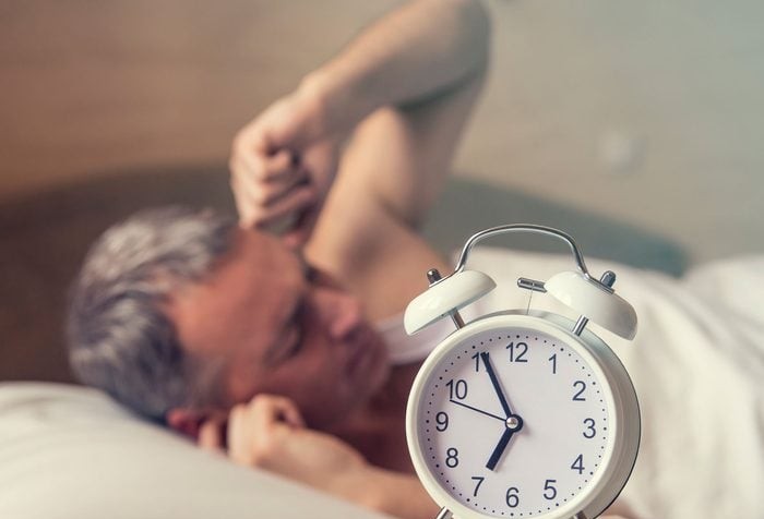 Muž v posteli s budíkom ukazujúcim šiestu hodinu ráno, pripravený na prebudenie a začatie dňa.