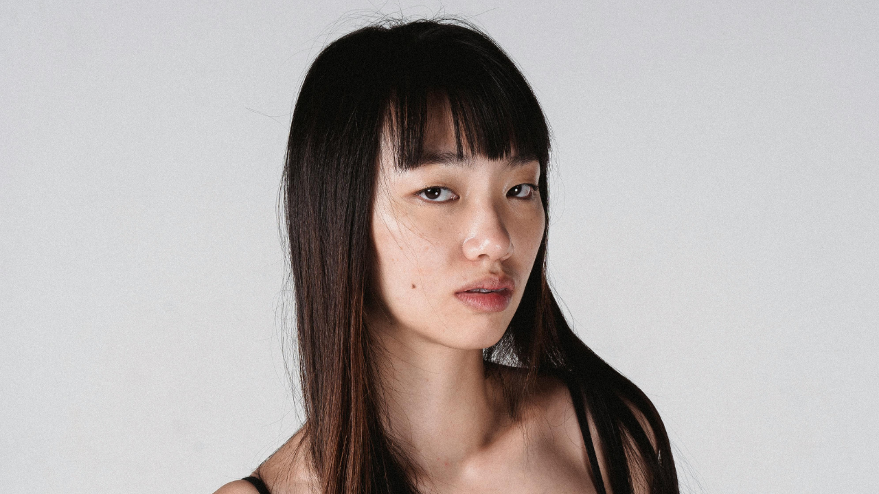 Mladá žena s dlhými vlasmi pozerá smerom do objektívu na neutrálnom pozadí.