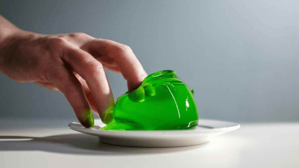 Ruka dotýkajúca sa zeleného želé na bielom tanieri v minimalistickom prostredí.