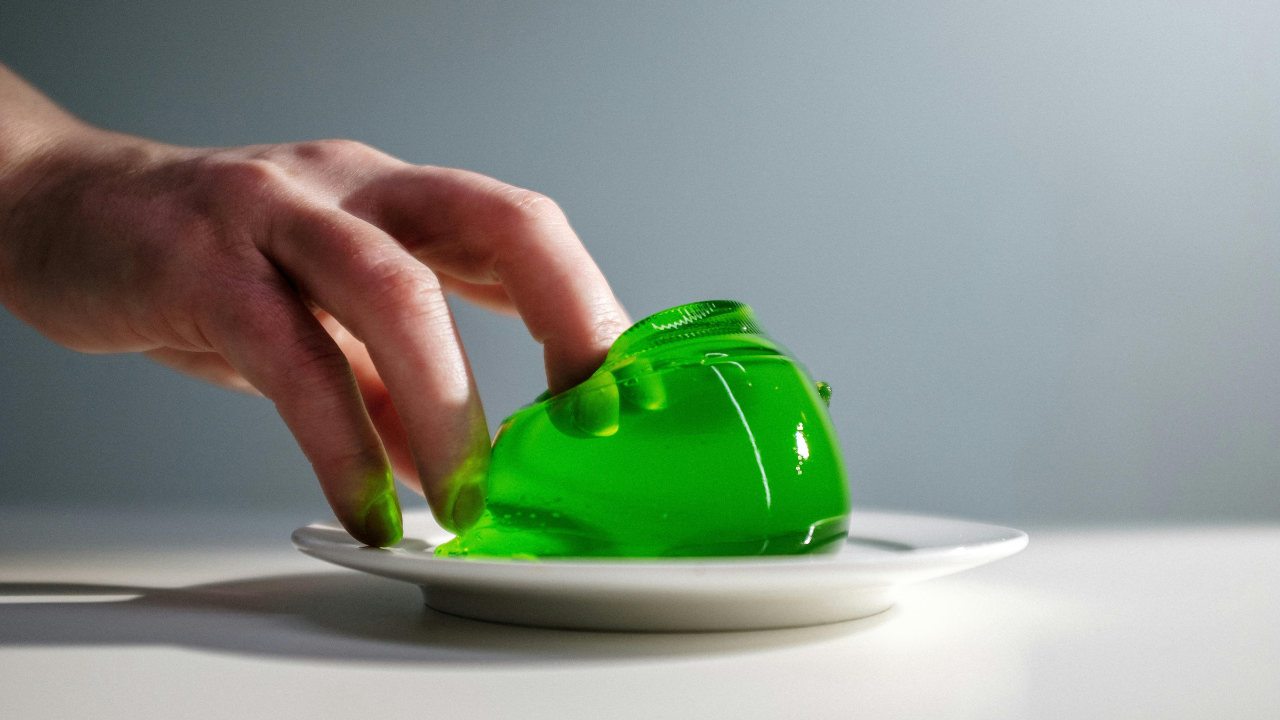 Ruka dotýkajúca sa zeleného želé na bielom tanieri v minimalistickom prostredí.