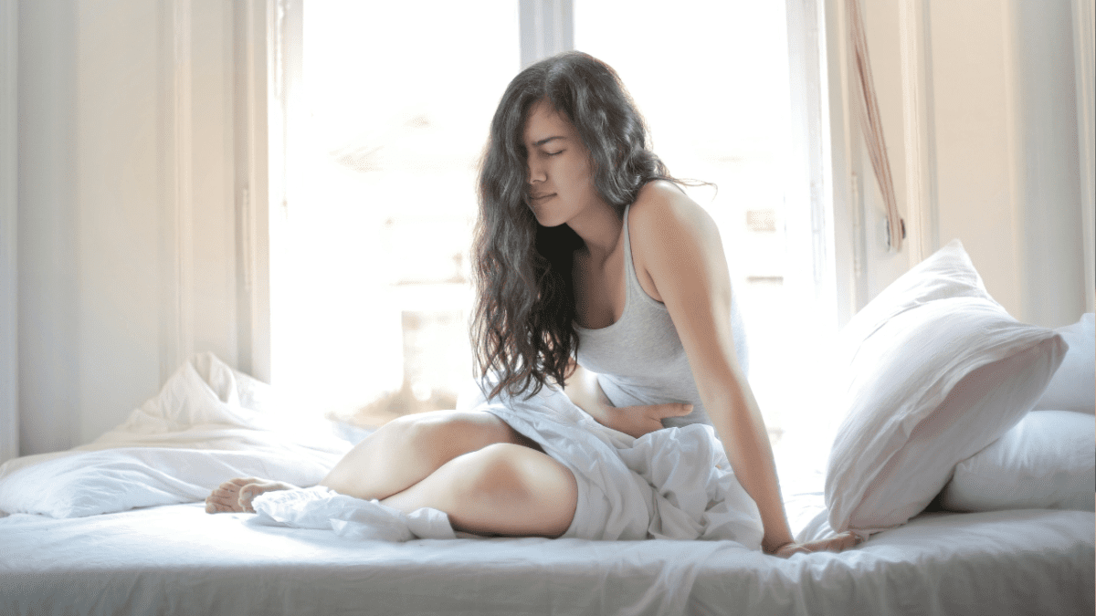 Žena so sklonenou hlavou sedí na posteli a drží si brucho, naznačujúc pocit bolesti alebo nepohodlia.