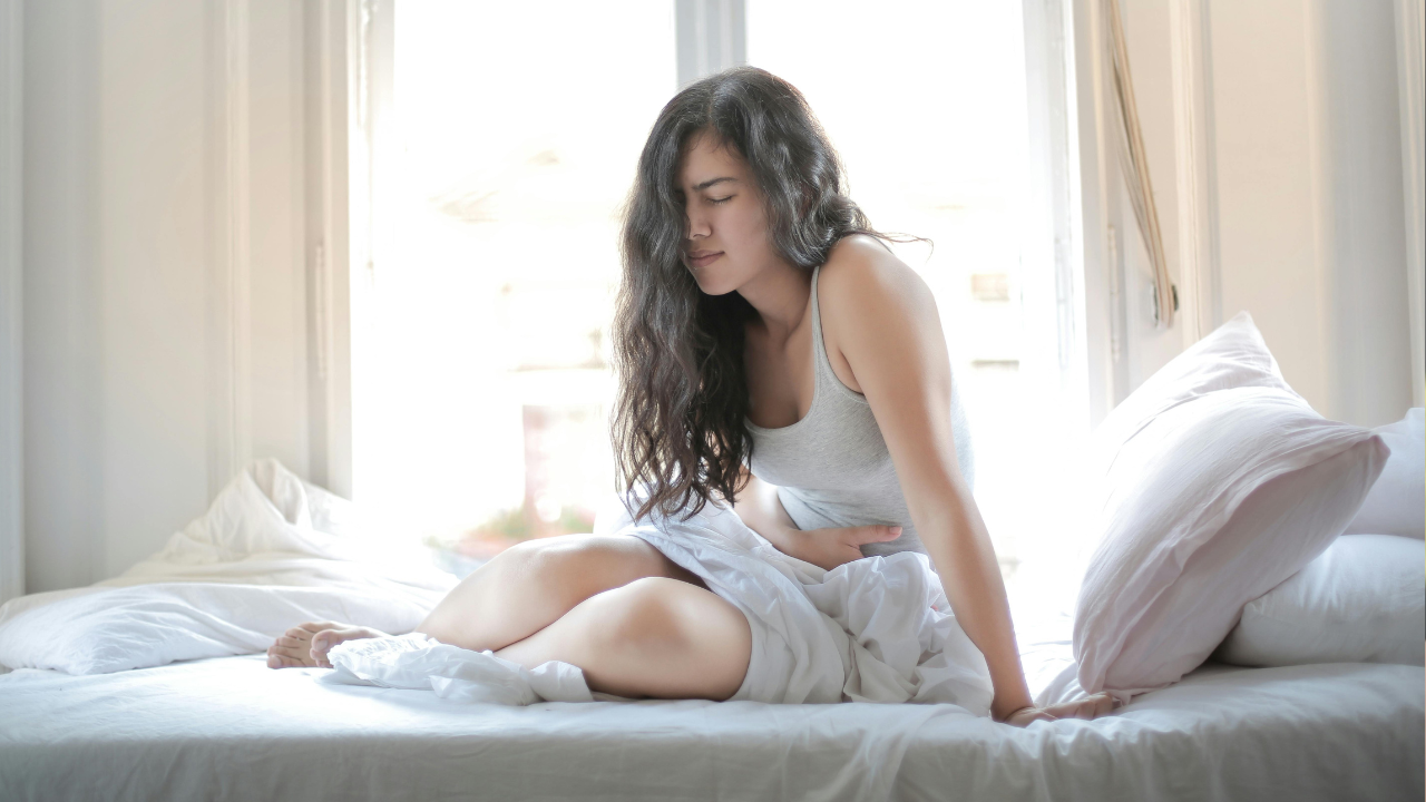 Žena so sklonenou hlavou sedí na posteli a drží si brucho, naznačujúc pocit bolesti alebo nepohodlia.