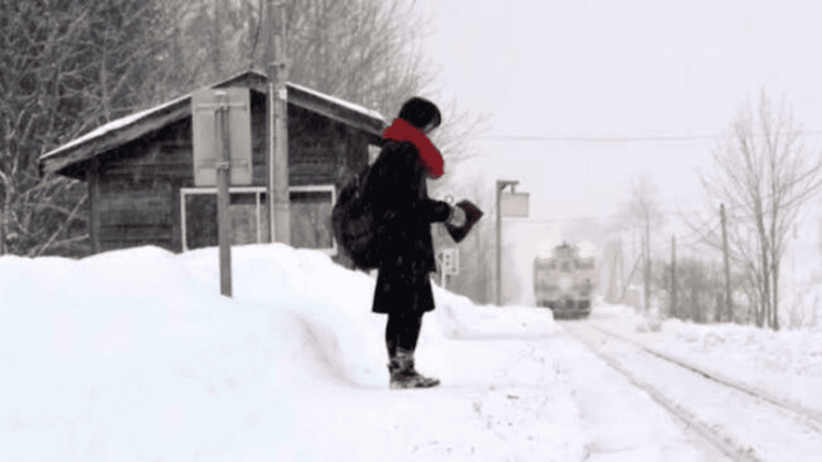 Osoba čaká na zasneženej stanici, blížiaci sa vlak v pozadí, zimná atmosféra.