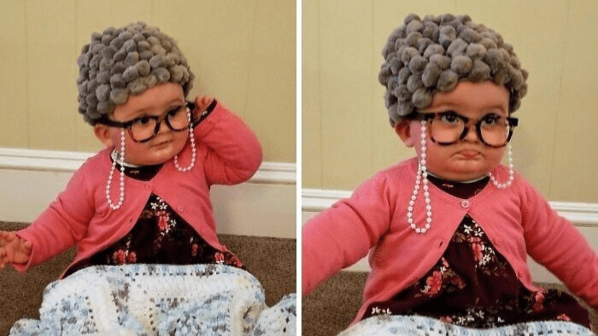 Roztomilé dieťa oblečené ako babička, s okuliarmi, šedou parochňou a ružovým svetrom, sedí na koberci.