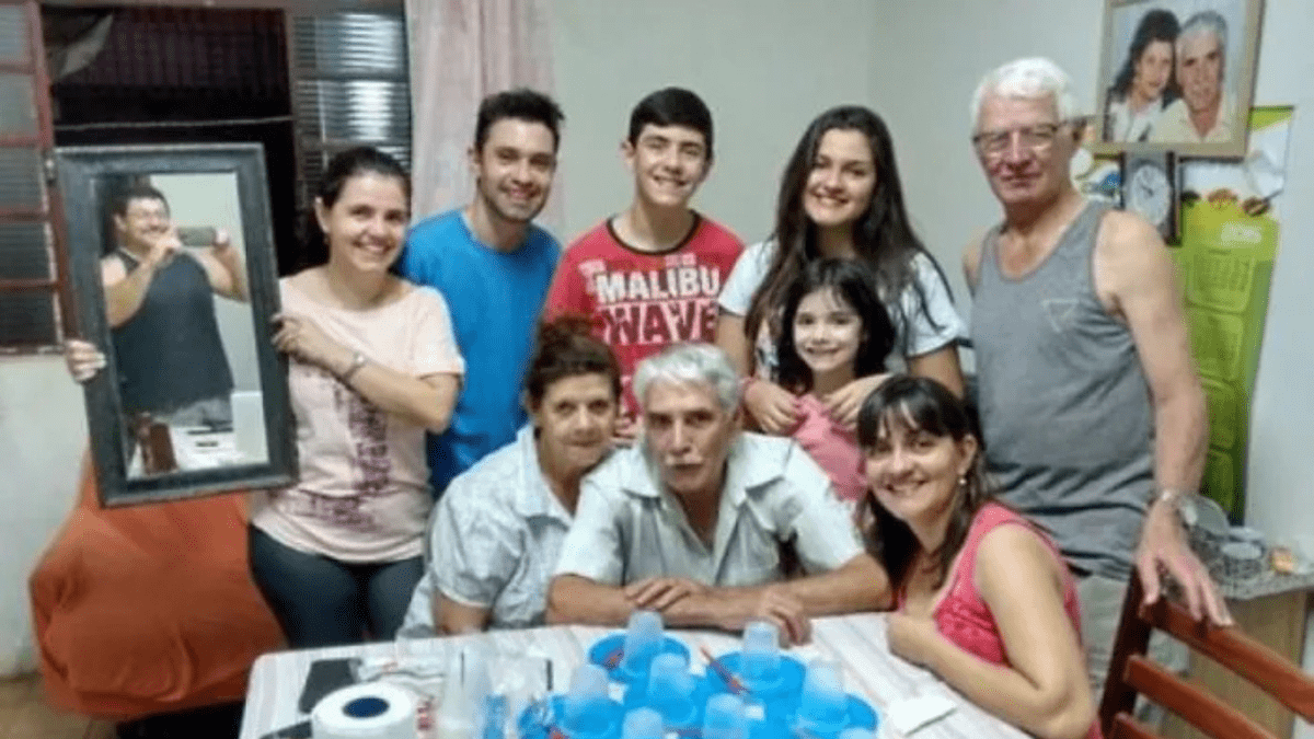 Skupinová rodinná fotografia s úsmevmi, žena drží zrkadlo, odraz muža viditeľný. Stôl s pohármi a drobnosťami.