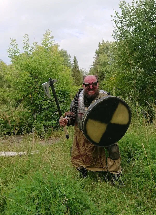 Bojovník v steampunk brnení s okuliarmi, sekerou a štítom stojí v tráve obklopený zeleňou.