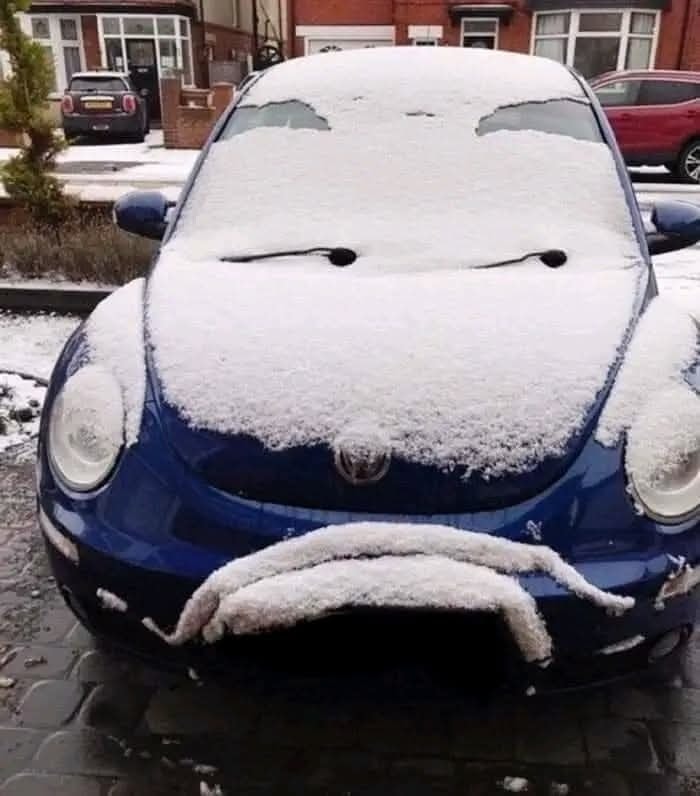 Modré auto pokryté snehom vyzerá ako tvár s očami a ústami, vďaka vrstve snehu na kapote a čelnom skle.