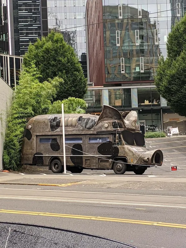 Autobus v tvare psa zaparkovaný v mestskom prostredí obklopený modernými budovami a zeleňou.