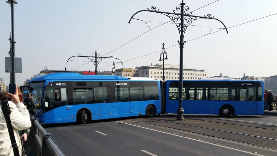 Modrý kĺbový autobus v strede cesty prechádza pod historickou lampou v mestskom prostredí.