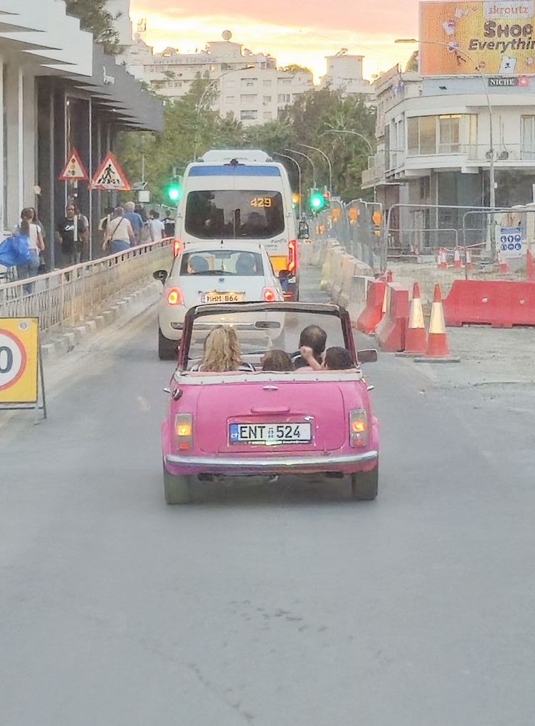 Ružové auto na večernej ulici, obklopené výstražnými značkami a dopravnými kužeľmi.