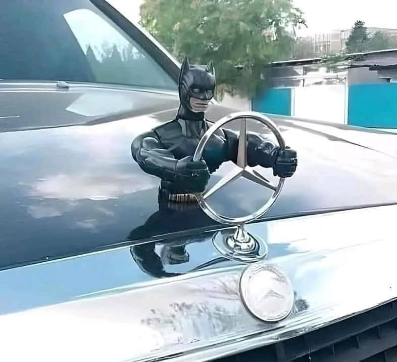 Figúrka superhrdinu s volantom Mercedesu na kapote auta, humorná montáž.