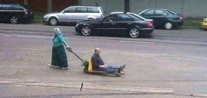 Žena ťahá muža na autecku po ceste vedľa zaparkovaných áut v meste.