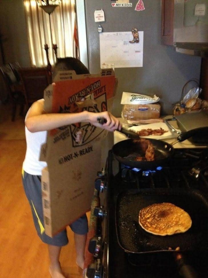 Osoba chrániaca sa pizzovou škatuľou pri varení slaniny a palacinky na panvici v kuchyni.