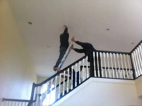 Dvaja muži opravujú strop na schodisku s použitím nebezpečne umiestnenej rebríka.