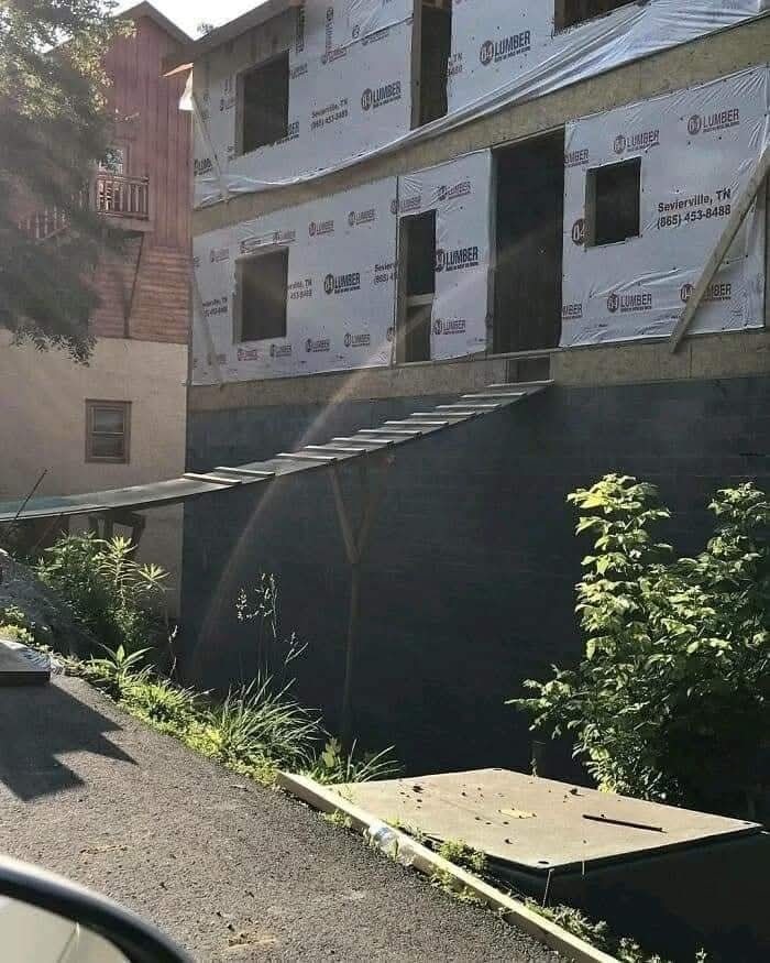 Stavebný dom s dočasným dreveným mostíkom cez priepasť, obklopený zeleňou.