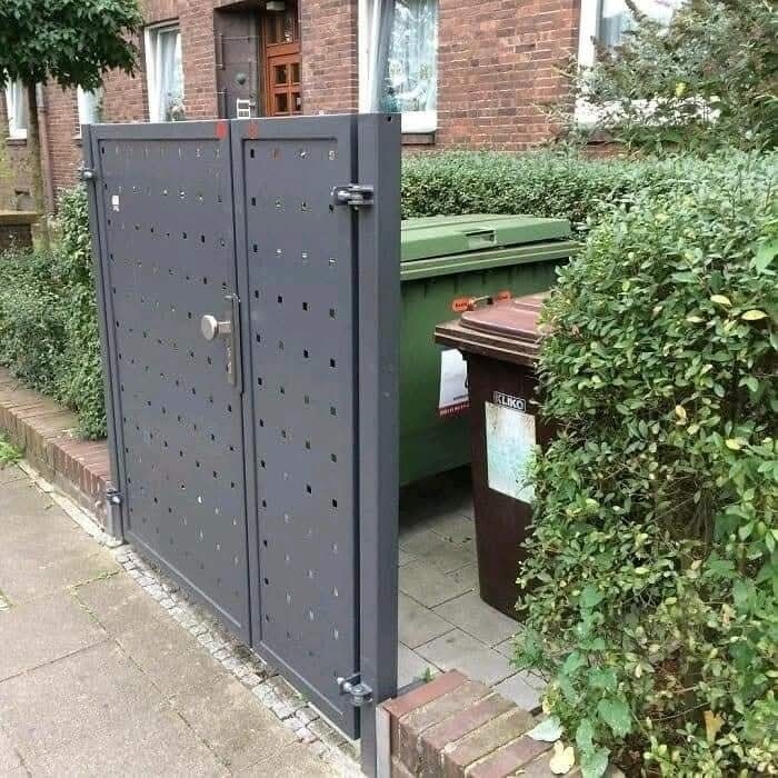 Dvojité brány s geometrickým vzorom pred kontajnerom na odpadky v mestskom prostredí.