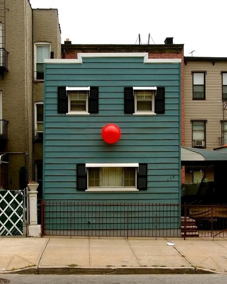 Modrý dom s červenou guľou pripomínajúcou nos na fasáde, vytvára ilúziu tváre v mestskej zástavbe.
