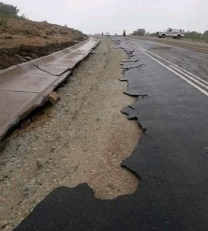 Rozpadávajúca sa asfaltová cesta, poškodený povrch vedľa krajnice, s autom a ľuďmi v diaľke na mokrom dni.