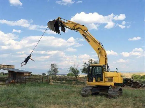 Stavebný stroj s človekom na lane, kreatívne využitie bagra v exteriéri pod modrou oblohou.
