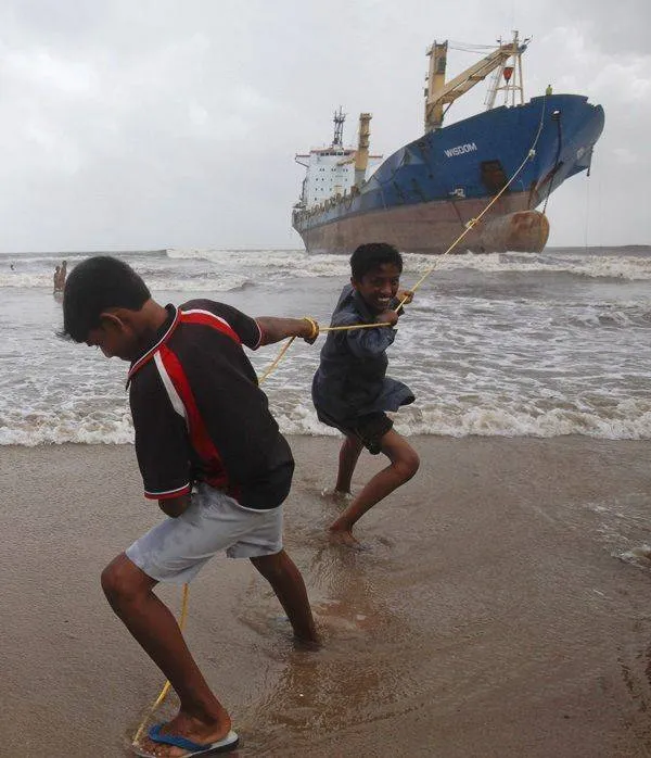 Dva deti na pláži ťahajú lano s veľkou loďou na pozadí, pri búrkovom mori.