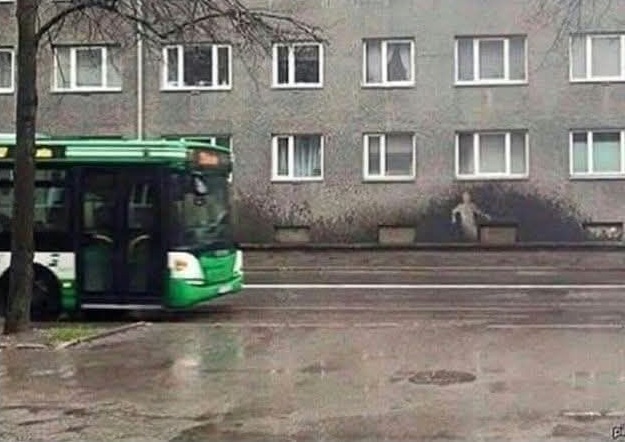 Autobus na ceste v daždivom počasí s mokrým chodníkom a stenou budovy v pozadí.