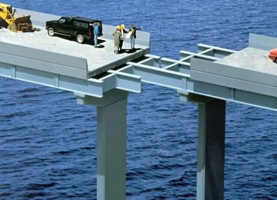 Nedokončený most ponad vodnú plochu s autami a pracovníkmi v bezpečnostných vestách diskutujúcimi na konci stavby.