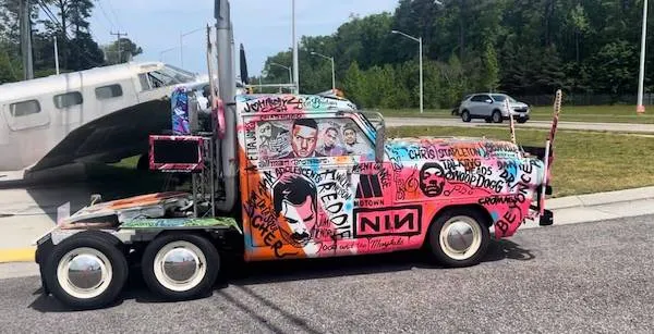 Farebný kamión s graffiti stojí pri ceste, v pozadí je lietadlo a zeleň.