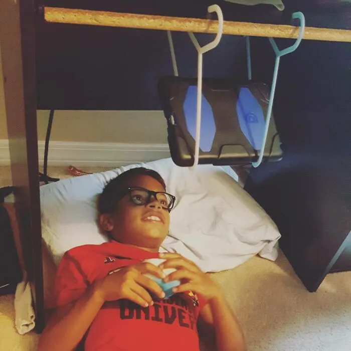 Dieťa leží na poduške a pozerá na tablet zavesený pod stolom.