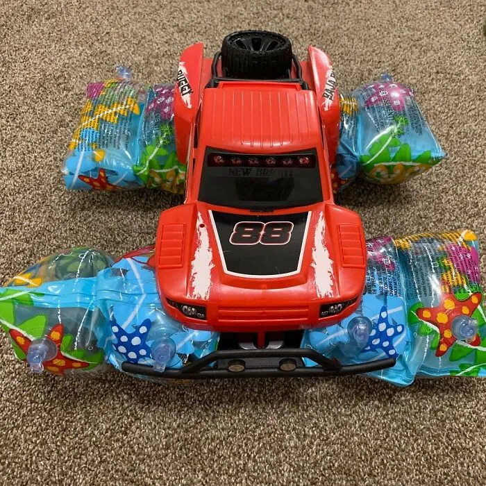 Červené auto hračky s balónovými rukávníkmi, umiestnené na hnedej koberci.