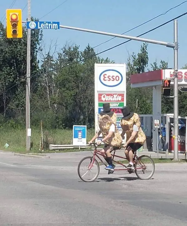 Dvojica v kostýmoch na tandemovom bicykli prechádza pri benzínke Esso pod značkou Leitrim.