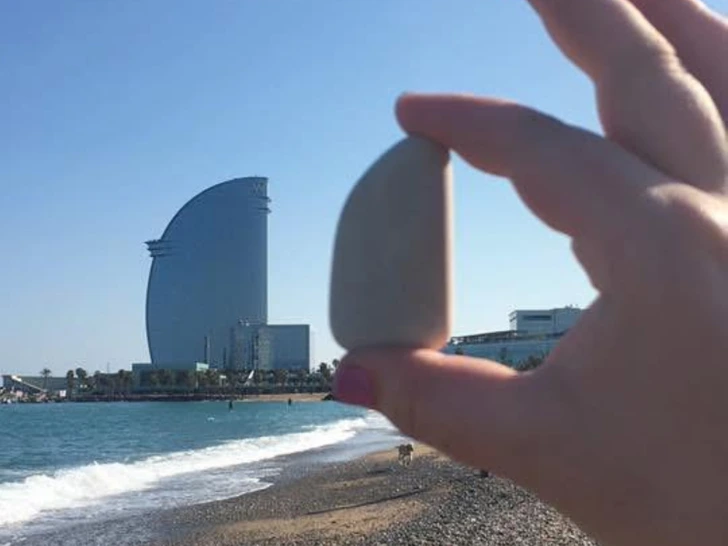 Ruka držiaca kameň pred plážou v Barcelone napodobňuje tvar budovy na horizonte.