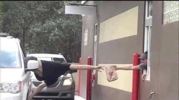 Muž v aute si berie jedlo z drive-thru okienka s extrémne natiahnutou rukou po tašku s nákupom.