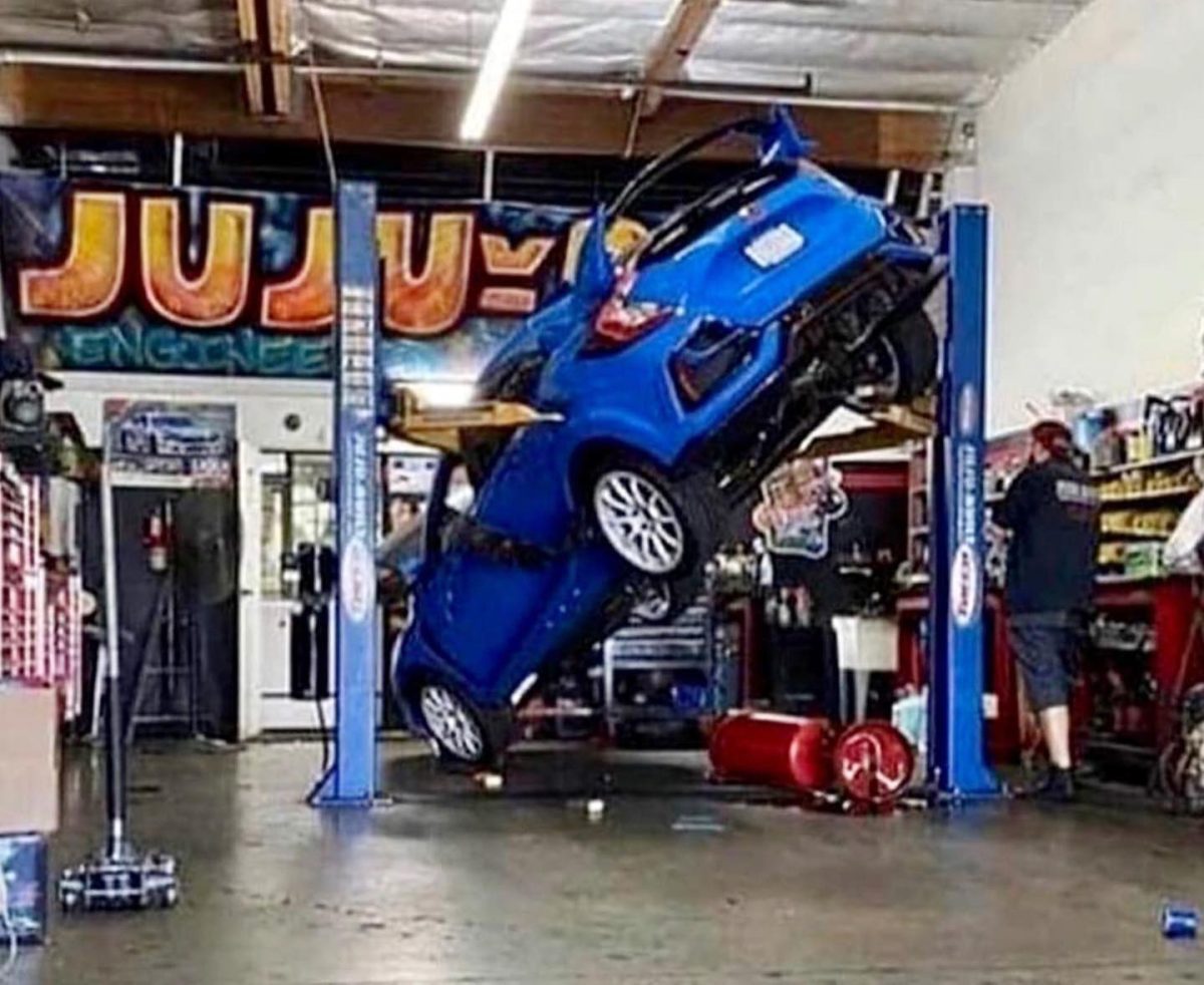 Auto neobvykle na zdviháku v autoservise, uvoľnený hasiaci prístroj, nástroje a vybavenie v pozadí.