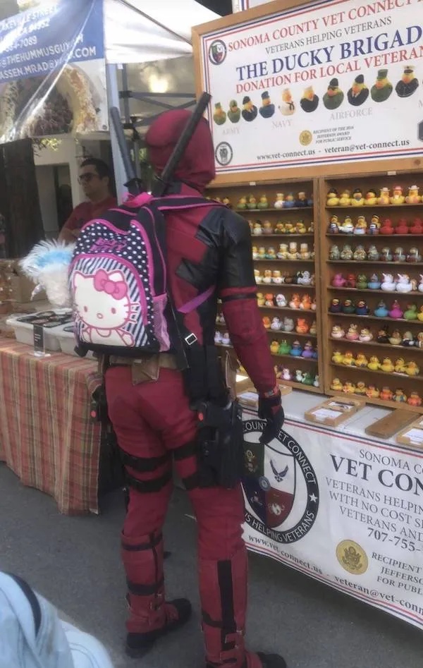 Superhrdina s ružovým ruksakom pri stánku s kačkami na podporu veteránov v outdoorovom prostredí.