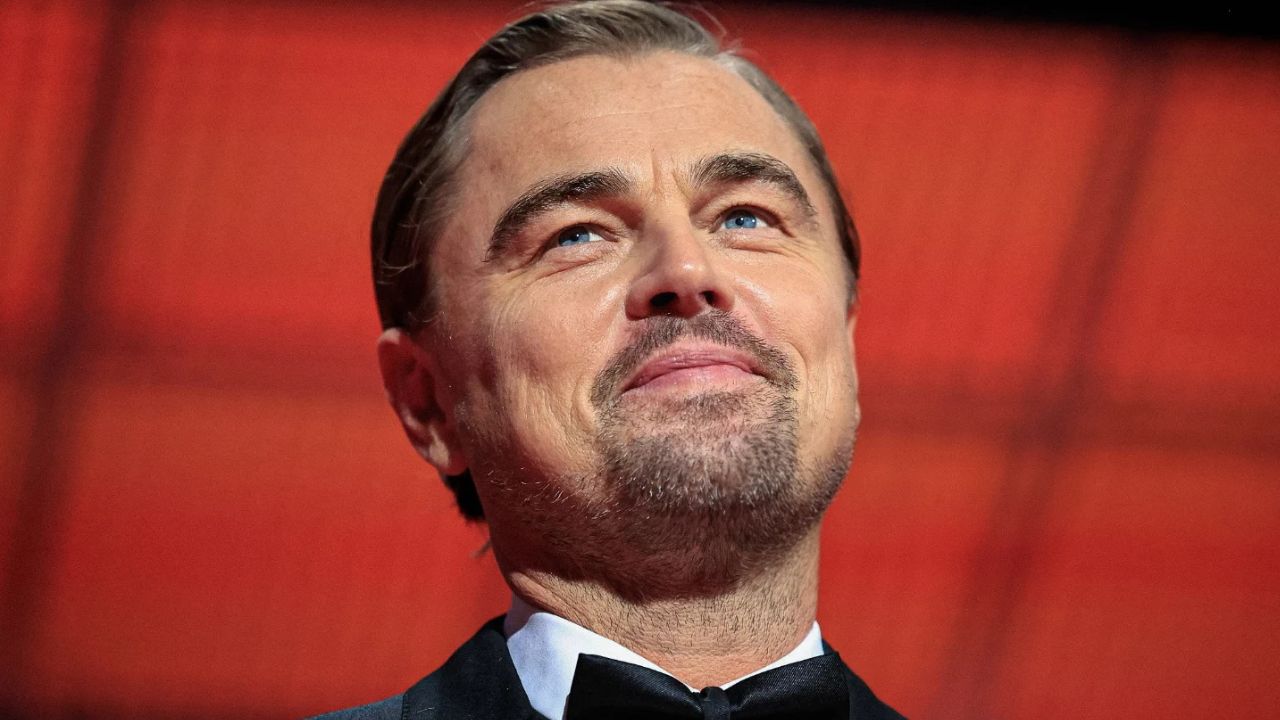 Osoba na červenom pozadí sa usmieva a má elegantný oblek a kravatu.