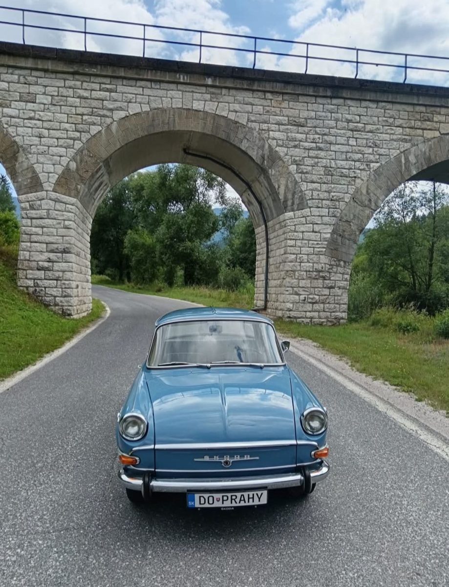 Modré retro auto na ceste pod kamenným viaduktom, obklopené zelenou krajinou a modrou oblohou.