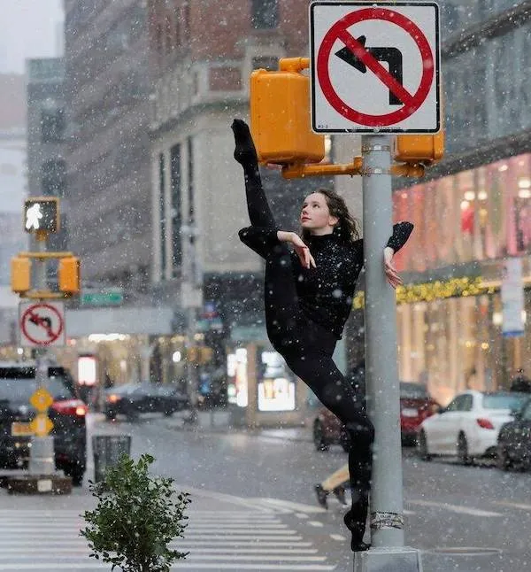 Baletka cvičí na zasneženom chodníku, opretá o semafor, počas zimného dňa v meste.