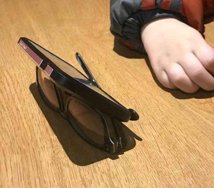 Mobilný telefón položený na okuliaroch vedľa detskej ruky na drevenom stole.