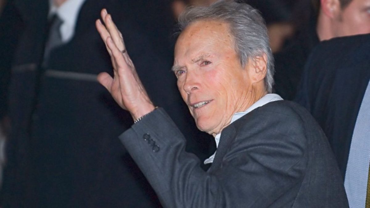 Osoba v tmavom obleku na večernej akcii s úsmevom a zdvihnutou rukou vo veľkom privítaní alebo pozdrave.