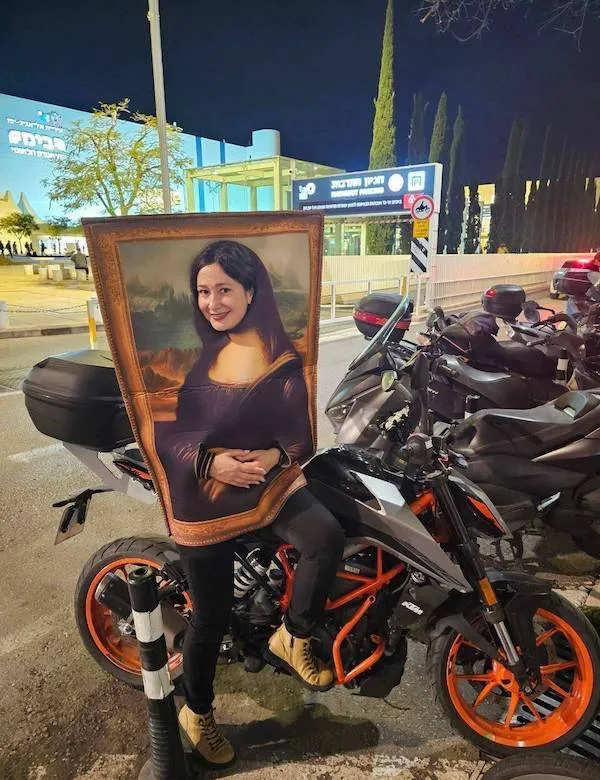 Osoba s kostýmom Mony Lisy sedí na motorke, nočná ulica v pozadí.