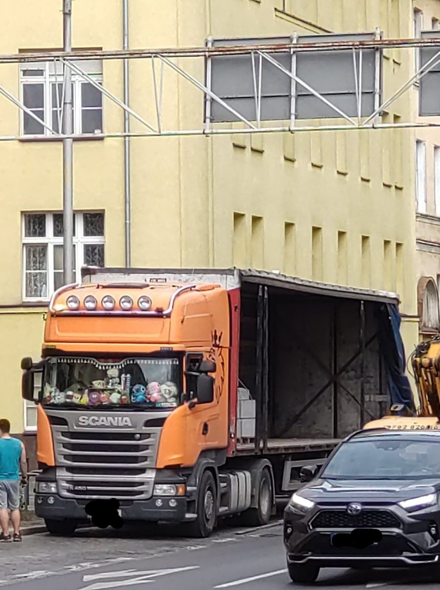 Oranžový kamión Scania parkujúci na ulici s otvoreným návesom, vedľa stojí auto a muž.