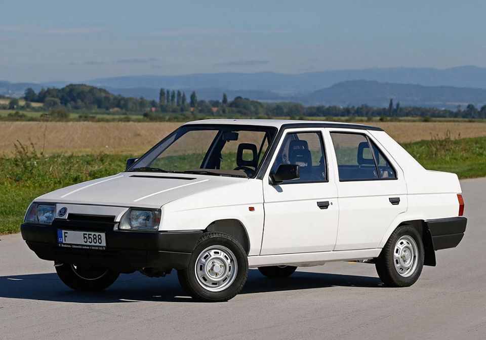 Biele retro auto na vidieckej ceste s modrou oblohou a kopcami v pozadí.