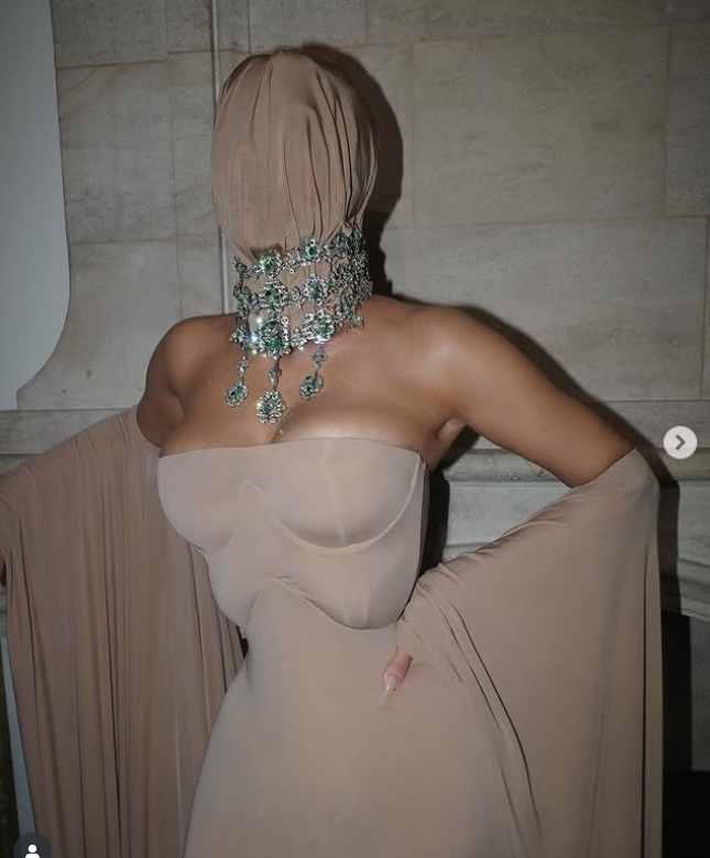 Osoba v telových šatách s krytím tváre a výrazným náhrdelníkom, stojaca pred svetlou stenou.