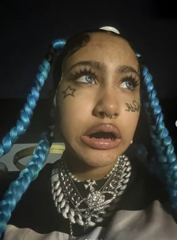 Osoba s modrými vrkôčikmi, výraznými šperkami a tetovaniami na tvári, v tmavom prostredí.