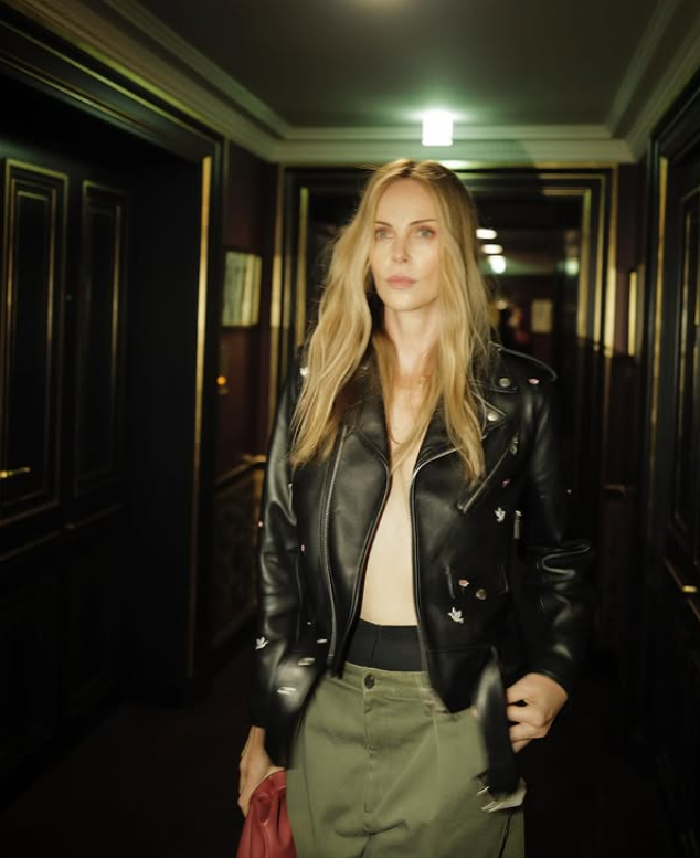 Žena v koženej bunde kráča chodbou s jemným osvetlením, drží červenú kabelku, luxusné prostredie.