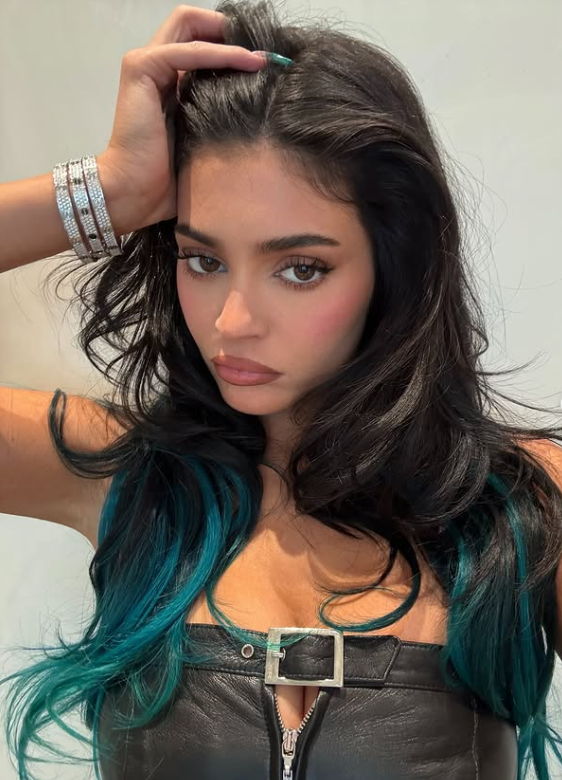 Tmavovlasá žena s tyrkysovými prameňmi vlasov a koženým korzetom, pózuje pri svetlom pozadí.