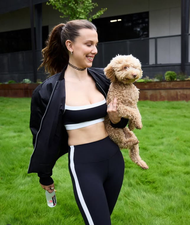 Žena v športovom oblečení drží malého psa na ruke, usmievajúc sa na trávnatom pozadí.