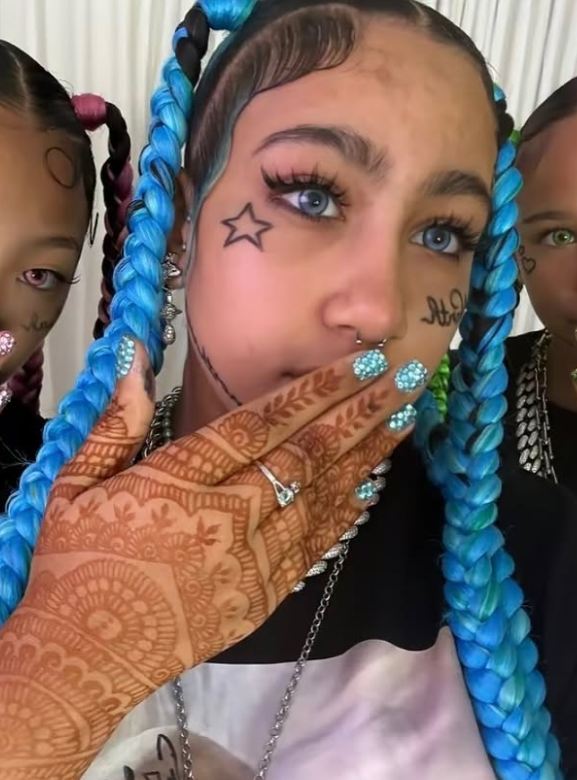 Osoba s modrými vrkočmi, hviezdičkou na tvári a zdobenými nechtami si zakrýva ústa hennou potetovanou rukou.