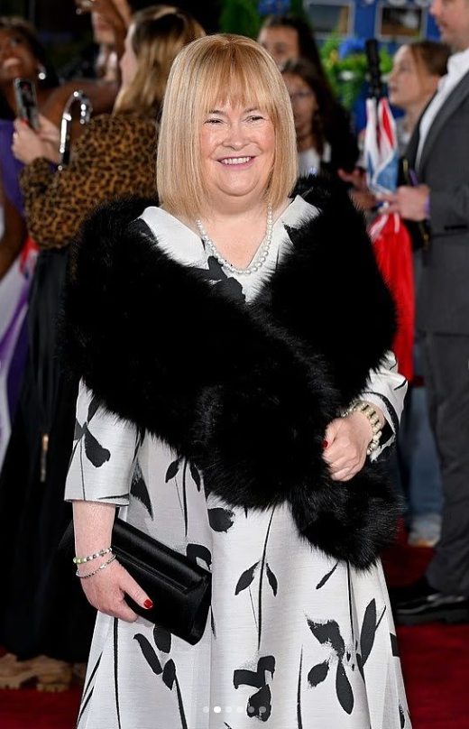 Prominentná žena v elegantných šatách a čiernom šále na spoločenskej udalosti, usmievajúca sa na červenom koberci.